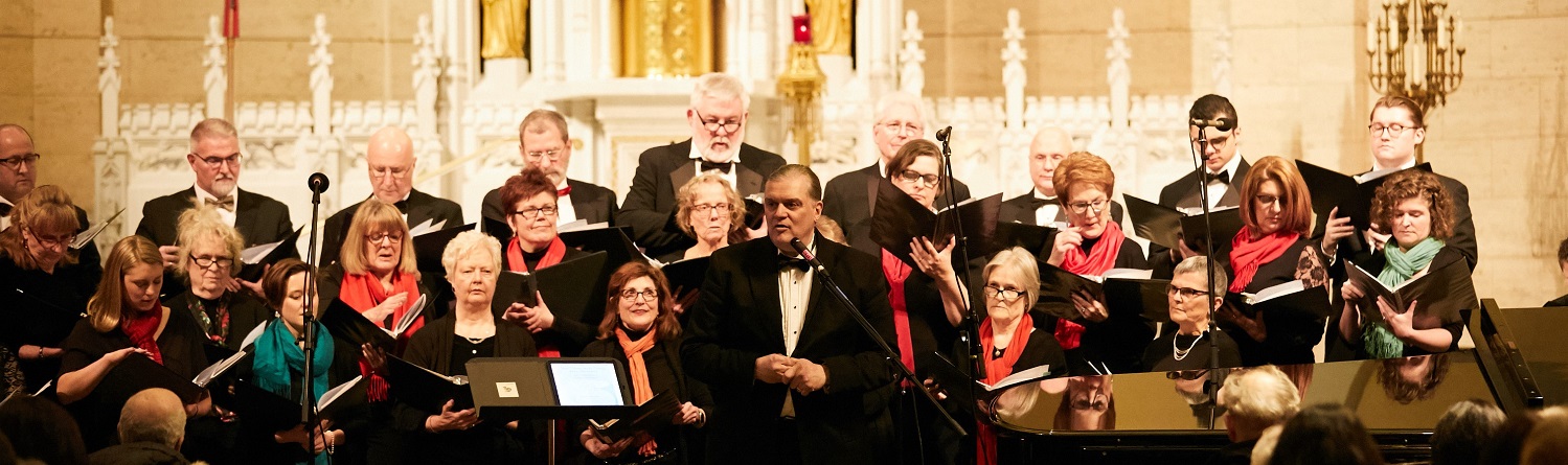 The group of singers lined up in three rows holding music books with conductor Philip in the middle-front of the image.